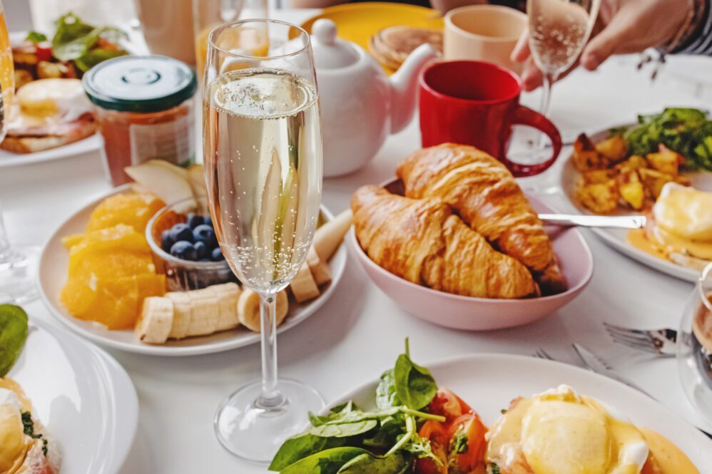 Glass of champagne close-up on the table with different breakfast and brunch dishes like croissants, eggs benedict, coffee and fruits. Selective focus