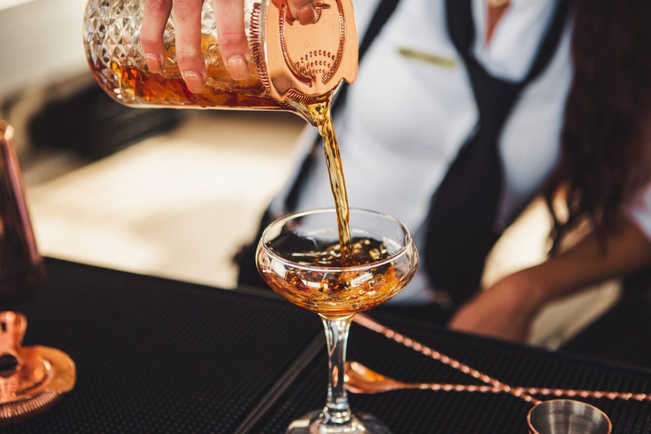A bartender in a white shirt and black tie pours a cocktail from a glass shaker through a strainer into a coupe glass on a bar counter.