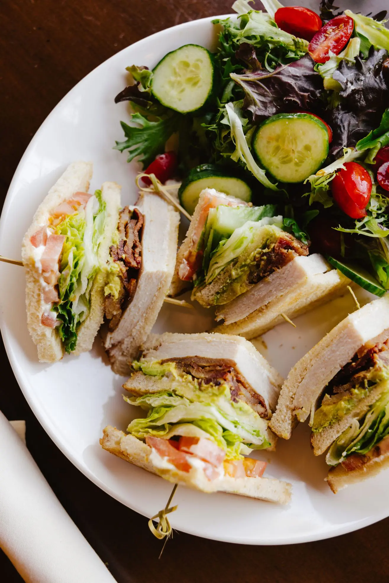 Club sandwich cut into fours with a side salad with cucumber and tomatoes on a white plate.