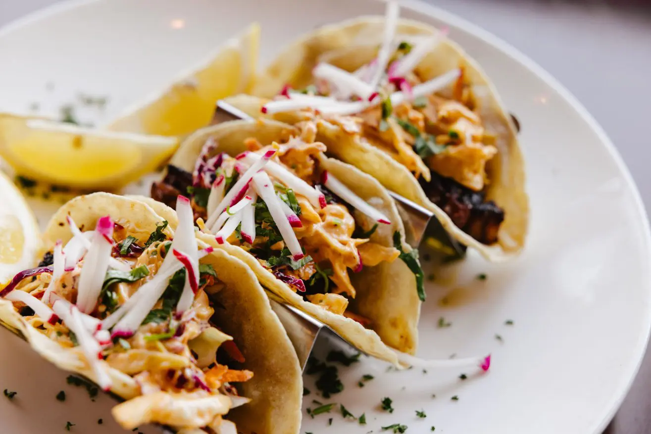 Three fish tacos on a white plate topped with sliced radish and cilantro.