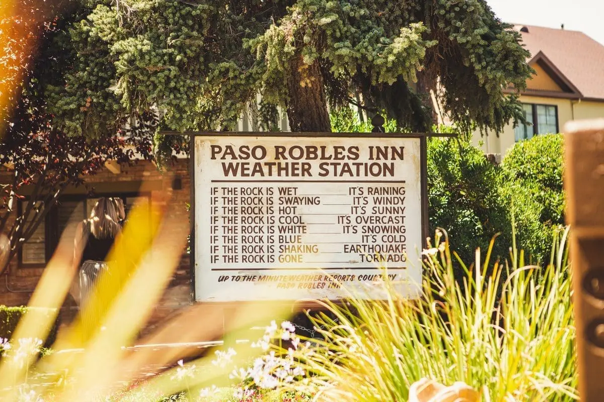 Rustic wooden sign reading "Paso Robles Inn Weather Station," surrounded by lush greenery, with a charming brick pathway in the background.