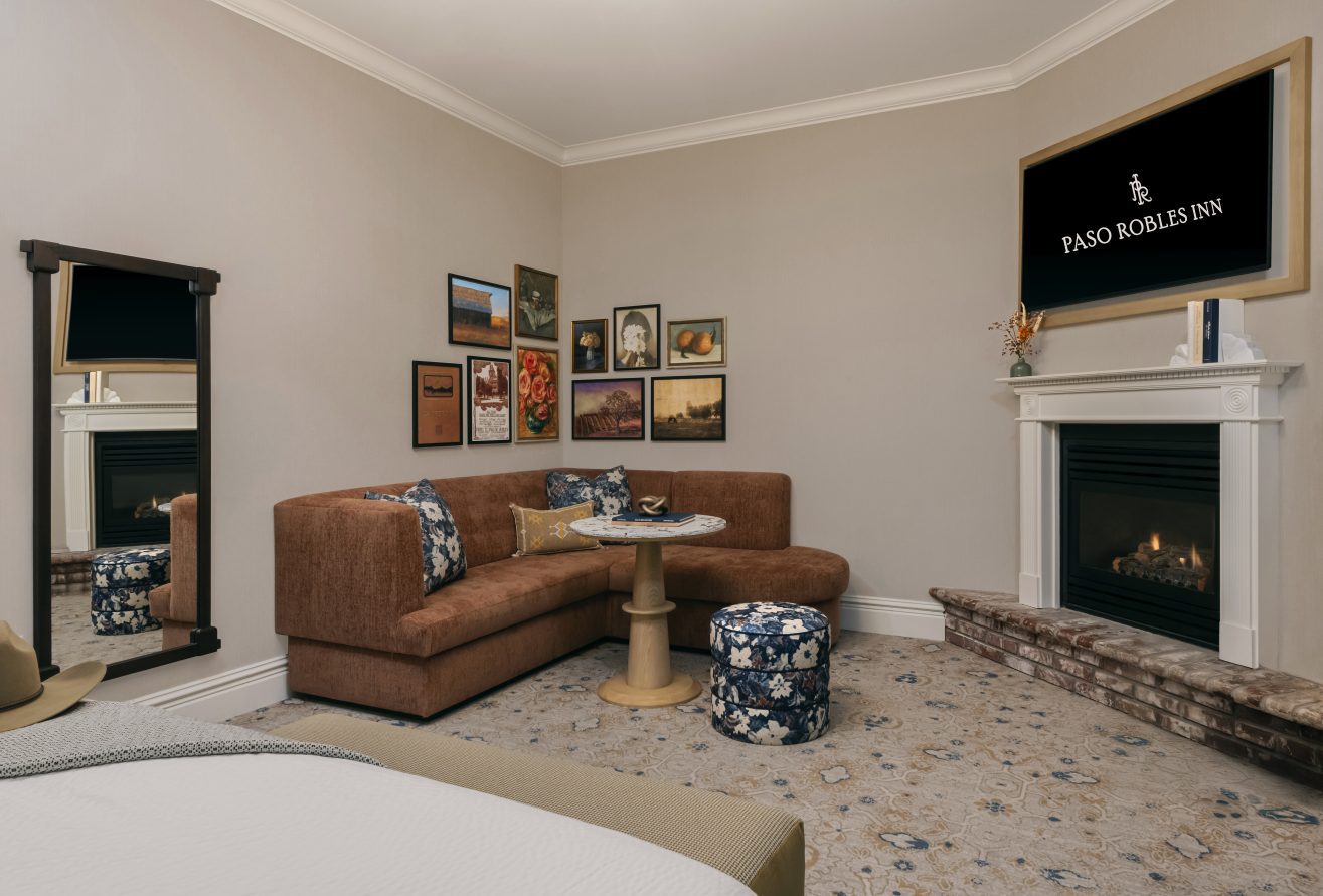 A cozy hotel room with a brown sectional sofa, a small round table, two blue floral ottomans, a wall-mounted TV above a fireplace, framed pictures on the wall, and a full-length mirror.