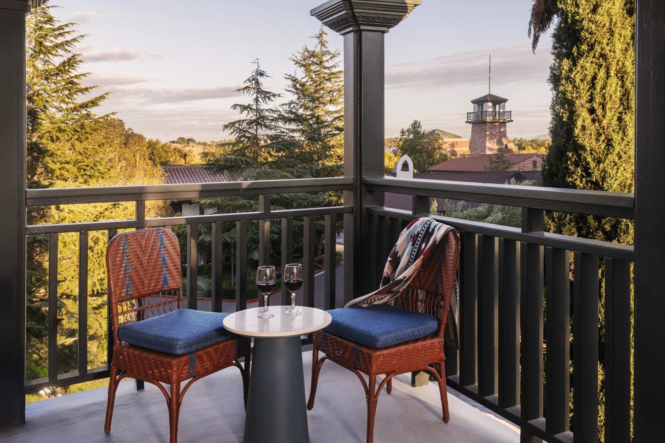 A cozy balcony with two wicker chairs and a small round table set with two glasses of wine, overlooking tall trees and a building with a clock tower at sunset.