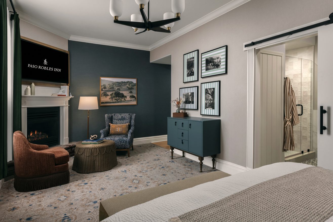 A cozy hotel room features a bed, fireplace, armchair with ottoman, a small blue dresser, framed wall art, and a sliding door leading to a modern bathroom. Warm lighting and neutral tones create a welcoming atmosphere.