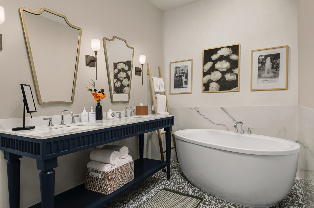 A modern bathroom with a blue double-sink vanity, two ornate mirrors, a freestanding bathtub, three framed pictures on the wall, and rolled white towels neatly stored below the counter.