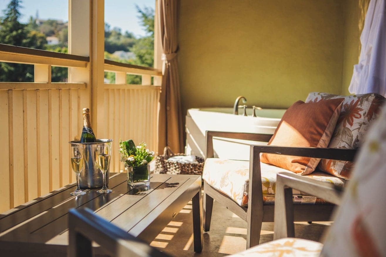 A sunlit balcony with cushioned chairs, a wooden table holding a wine bucket with champagne and glasses, a potted plant, and a bathtub in the corner. Trees and greenery are visible outside.