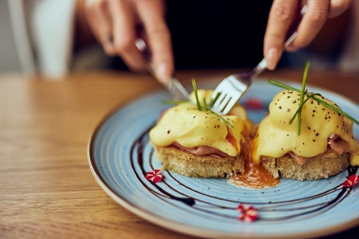A person cuts into eggs Benedict on a blue plate, with runny yolk, hollandaise sauce, ham, and chives on toasted bread.