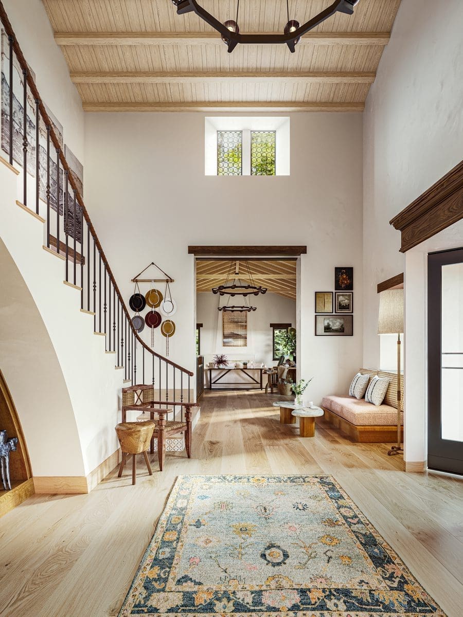 Bright entryway with light wood floors and vaulted ceiling, curved staircase with black railing, patterned rug, bench with cushions, and wall decor. Living area with wooden beams visible in the background.