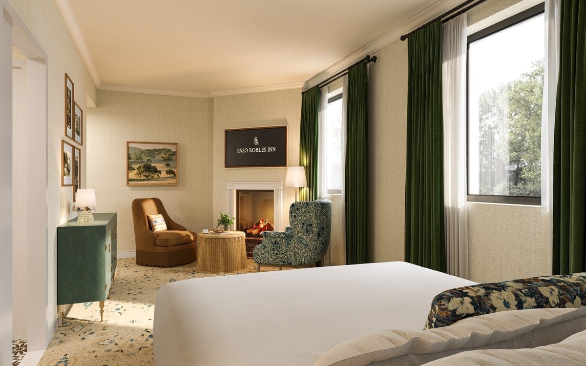 A cozy hotel room with large windows, green curtains, a bed in the foreground, an armchair and round ottoman by a fireplace, and framed artwork on the walls. Sunlight fills the softly decorated, inviting space.