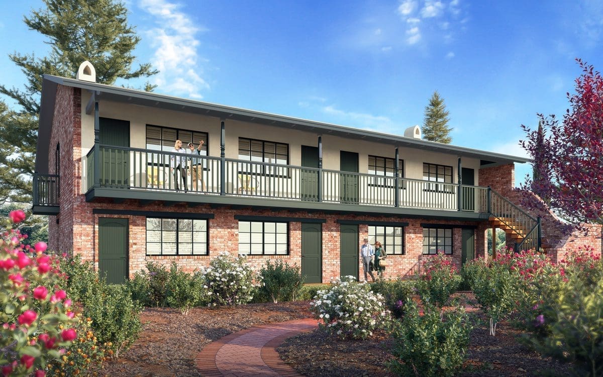 Two-story brick building with green railings and trim, surrounded by flowering bushes and trees, under a blue sky. Two people stand on the upper balcony, and one person stands near the ground-floor entrance.