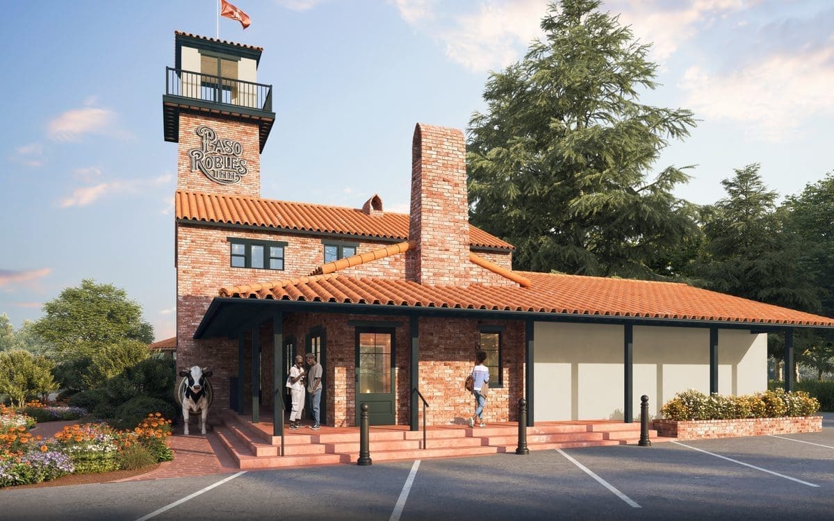A brick building with a red-tiled roof, chimney, and a tower with a sign reading Paso Robles Inn. People are at the entrance, and there are flowers, greenery, and a clear sky with soft clouds.