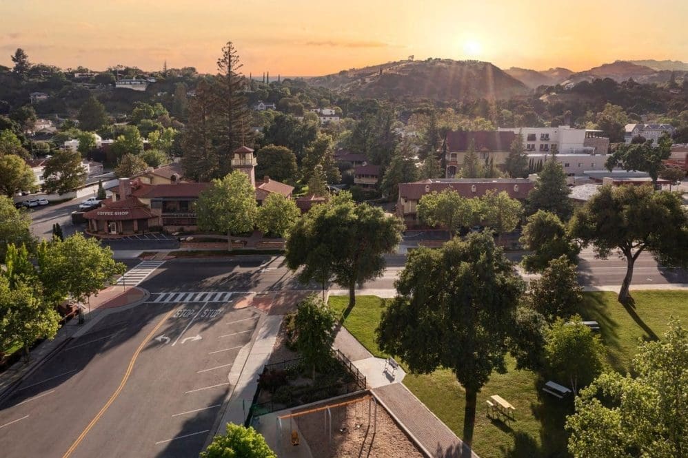 A tree-lined suburban street with buildings, parked cars, and a playground in the foreground, set against rolling hills and a vibrant sunset sky.