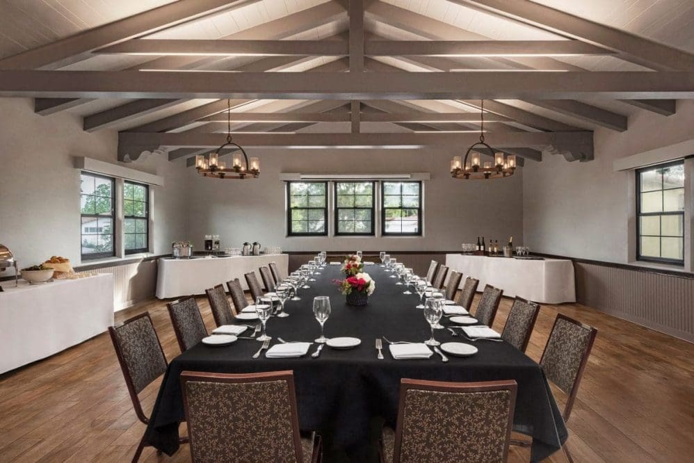 A formal dining room with a long table set for a meal, surrounded by chairs. The table has a black tablecloth, white plates, glasses, and a flower centerpiece. Large windows and chandeliers light the spacious, elegant room.
