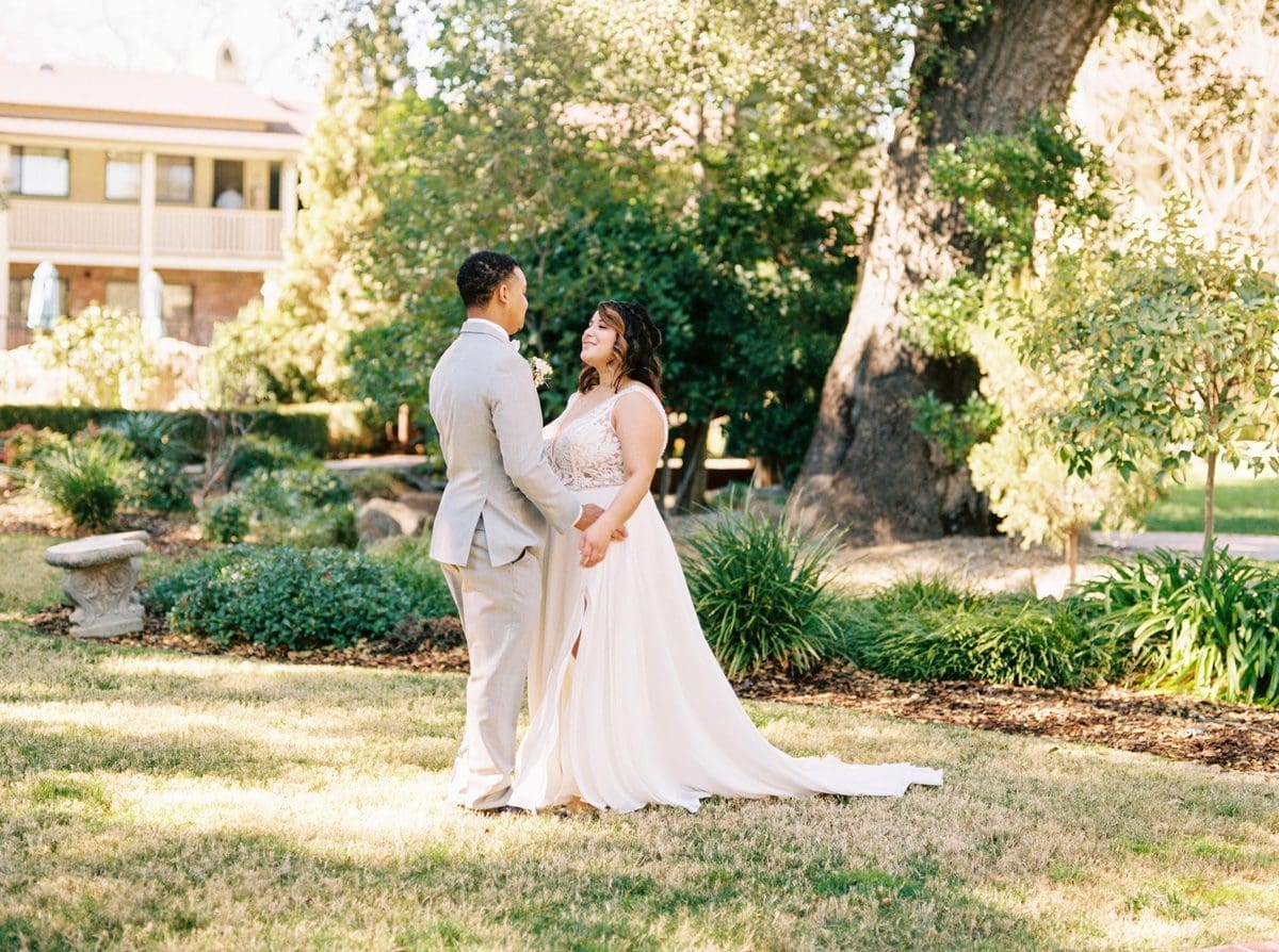 A bride and groom stand facing each other and smiling, holding hands in a sunlit garden with trees, greenery, and a large tree trunk in the background. The bride wears a white dress; the groom wears a light gray suit.