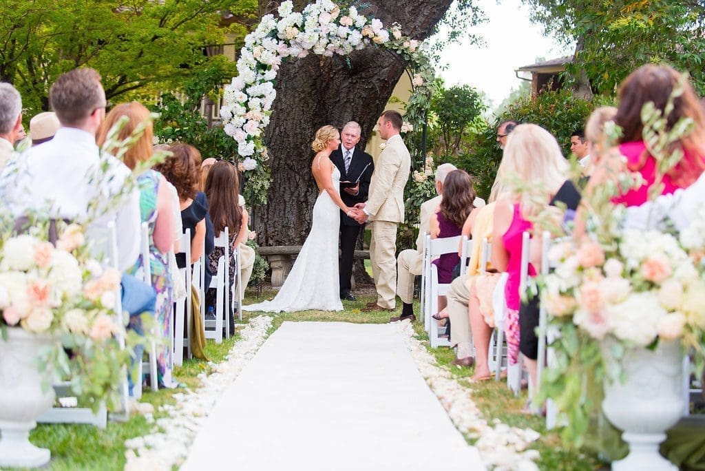 A bride and groom stand under a floral arch outdoors, holding hands and facing each other during their wedding ceremony, surrounded by seated guests on either side of a white aisle.