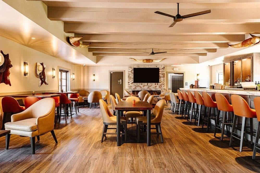 Spacious restaurant interior with wooden floors, beige and orange upholstered chairs, tables, a bar area with stools, wall-mounted bull horns, and a large TV above a brick fireplace—capturing the inviting charm of Paso Robles restaurants.