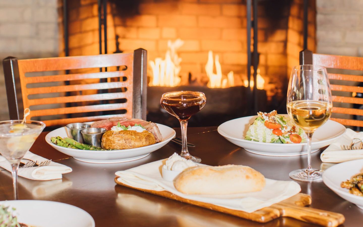 A cozy Steakhouse Paso Robles table set with plates of food, including a baked potato, steak, salad, and bread roll. Two wine glasses and a cocktail complete the scene, with a lit fireplace glowing in the background.