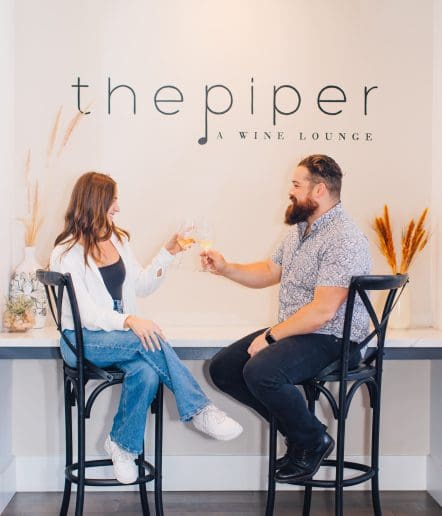A woman and man sit on tall chairs, clinking wine glasses and smiling at each other in front of a sign that reads the piper: A WINE LOUNGE—one of the coziest spots for Paso Robles Events. Dried plants decorate the shelf behind them.