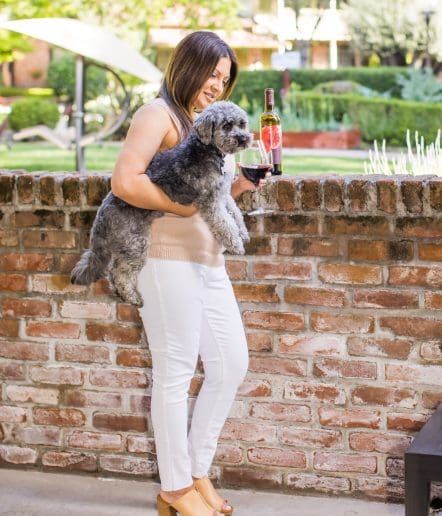 A woman in white pants and a beige top stands by a brick wall, holding a small gray dog. She smiles and holds a glass of red wine, with a bottle of wine on the wall. Greenery and a garden are visible in the background.