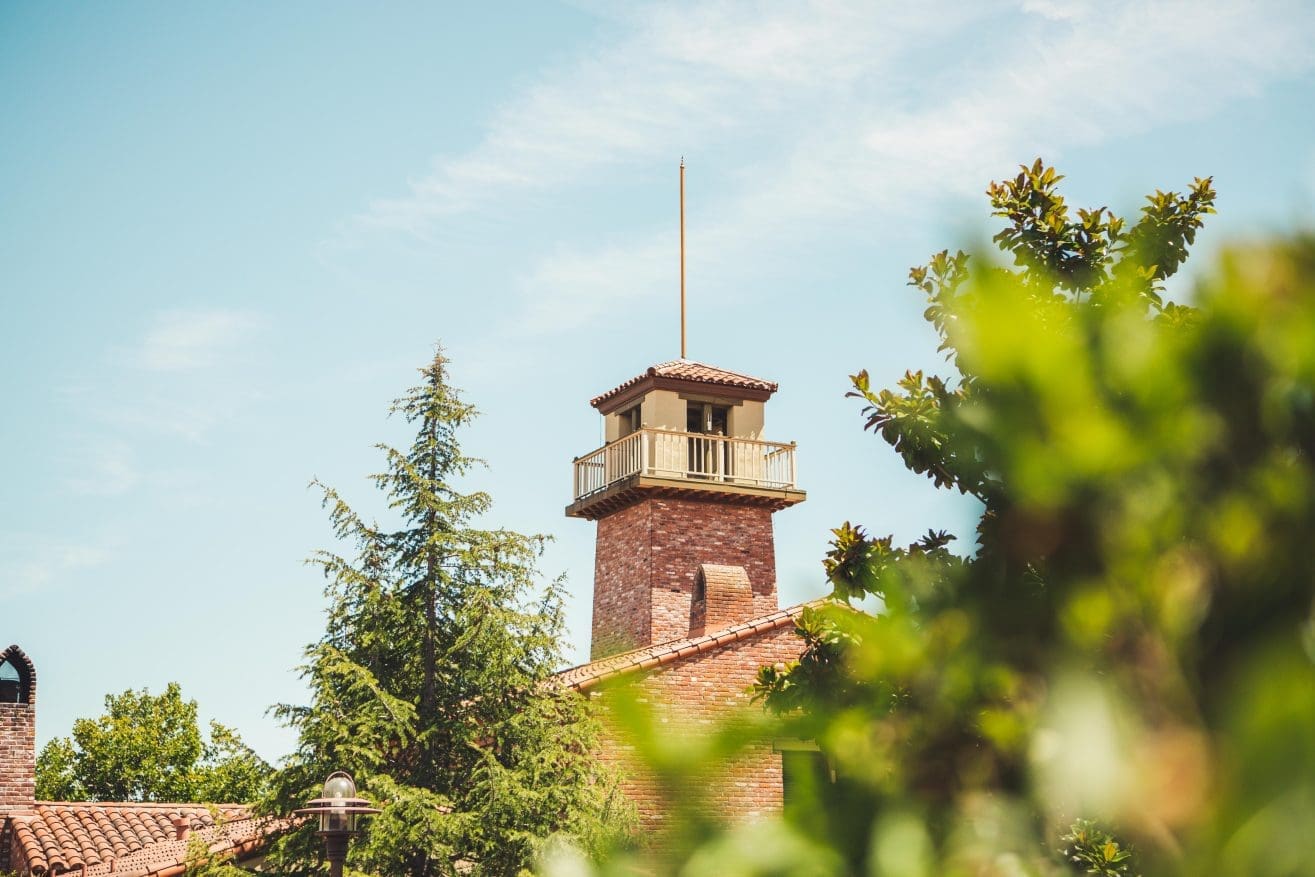A brick tower with a balcony and tiled roof rises above green trees and leafy branches under a bright blue sky.