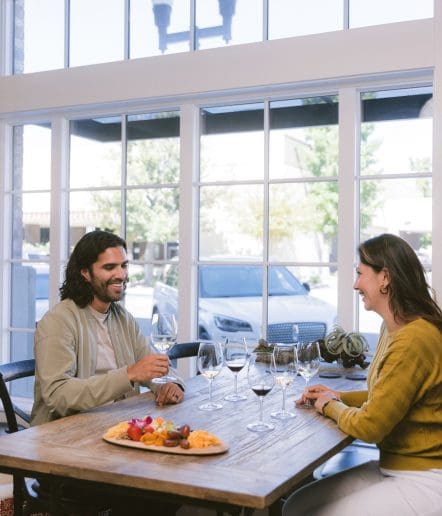 A man and a woman sit across from each other at a wooden table with wine glasses and a platter of cheese and fruit, smiling and talking in front of large windows during the day.