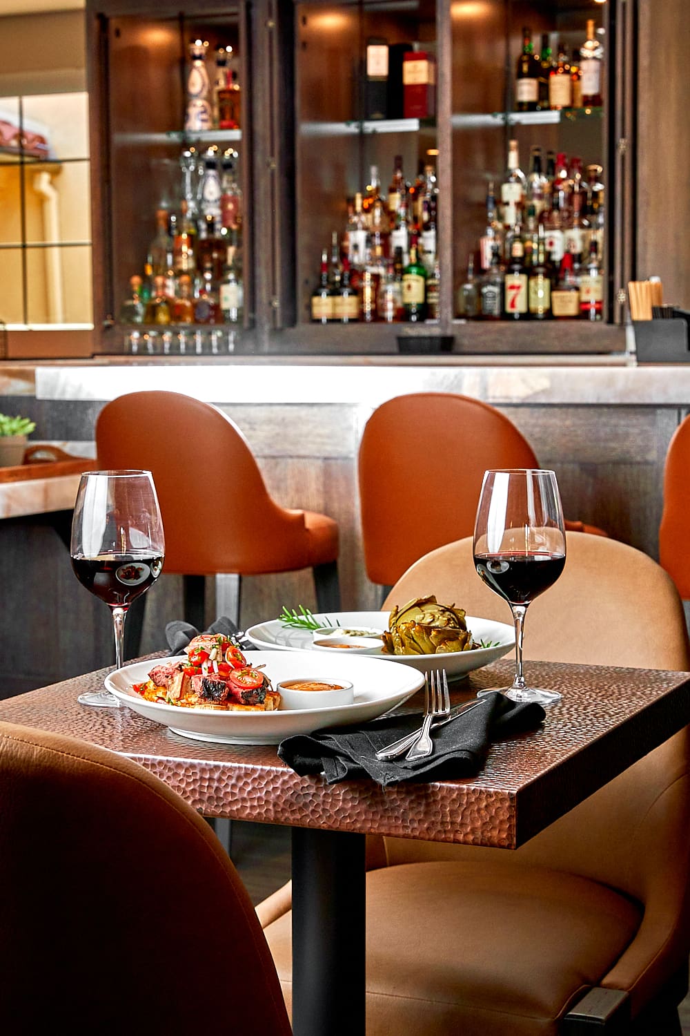 A restaurant table set for two with plates of gourmet food, two glasses of red wine, and black napkins, in front of the Cattlemen's Lounge Paso Robles bar stocked with bottles and brown leather chairs.