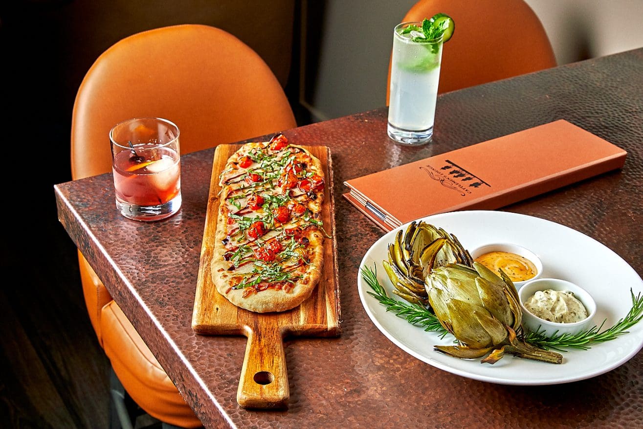 A restaurant table at Cattlemen's Lounge Paso Robles features flatbread with tomatoes and greens on a wooden board, grilled artichoke with sauces, two cocktails, an orange menu, and two partially visible orange chairs.
