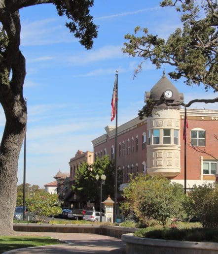 A historic brick building with a clock tower sits on a sunny street corner in Paso Robles, surrounded by trees and flags. Cars line the street, the sky is clear and blue—capturing the charm often celebrated during Paso Robles Events.