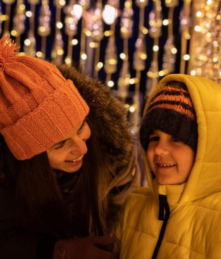 A woman in a pink knit hat and a child in a yellow puffer jacket and striped hat smile together, surrounded by warm, glowing lights and festive decorations—capturing the joyful spirit of Paso Robles Events.