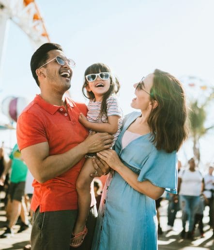 A smiling family of three wearing sunglasses enjoys a sunny day outdoors at Paso Robles Events. The father in a red shirt holds their young child, while the mother in a blue dress stands beside them with people and amusement rides in the background.