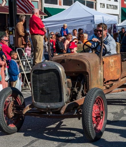 A man in sunglasses drives a rusty, vintage car with red wheels in a street parade at Paso Robles Events, while people and children watch and smile from the sidewalk under white tents.