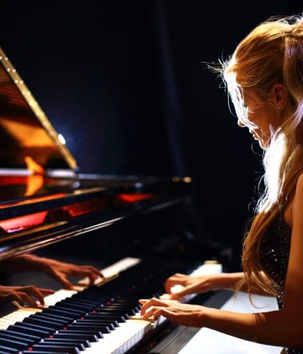 A woman with long blonde hair plays a grand piano under dramatic lighting at a Paso Robles Events venue, her reflection shimmering on the polished piano surface.