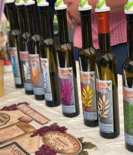 Several bottles of olive oil with colorful labels are arranged in a row on a table, accompanied by pamphlets or menus—perfect for tastings at Paso Robles Events. People are visible in the background, partially out of frame.