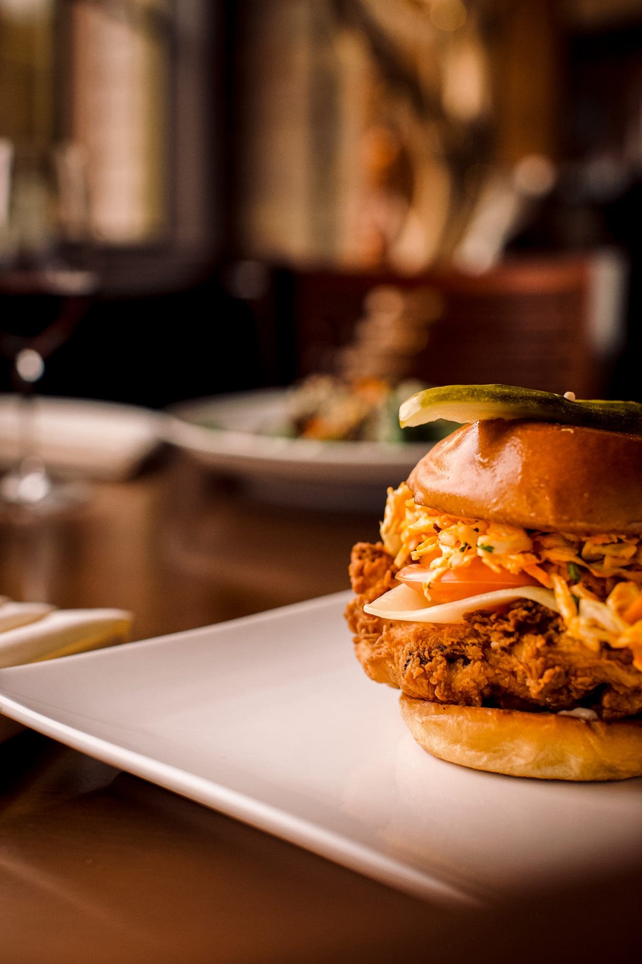 A close-up of a crispy fried chicken sandwich topped with coleslaw and pickles, served on a white plate in a warmly lit Steakhouse Paso Robles restaurant setting.