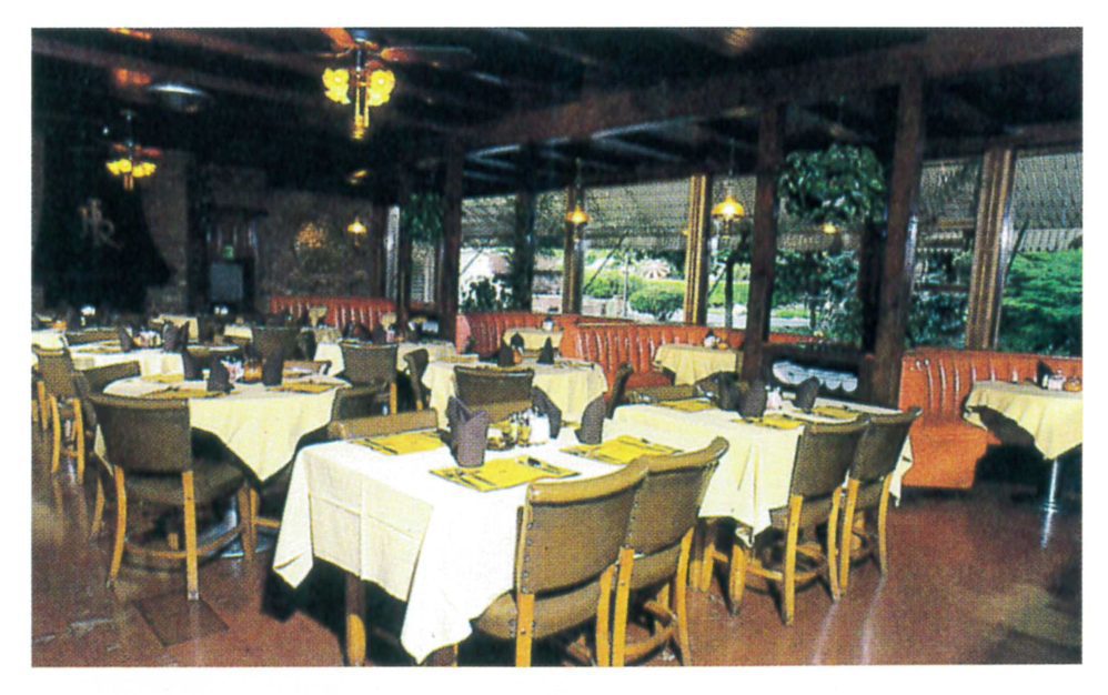 A restaurant dining area with tables covered in yellow cloths, brown chairs, and place settings. Large windows provide views of greenery outside, and orange booth seating lines the wall. Ceiling fans hang above.