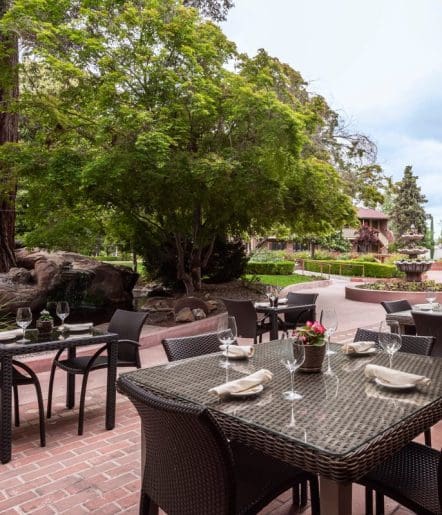 Outdoor patio dining area with wicker tables and chairs set with glassware and napkins, surrounded by lush green trees, manicured gardens, and a brick pathway in a serene setting.