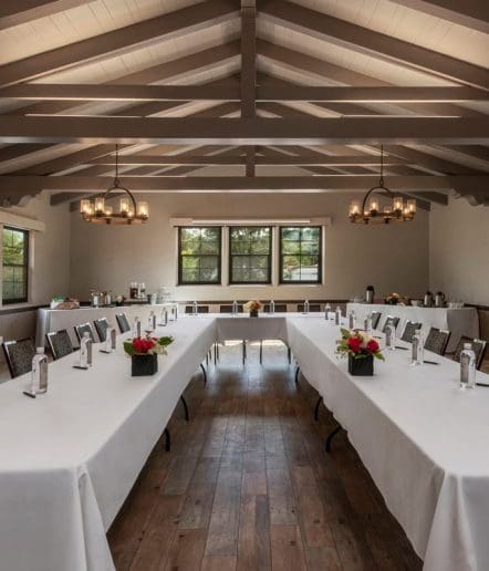A conference room with wooden floors and exposed beams, featuring two U-shaped tables covered with white tablecloths, floral centerpieces, water bottles, and chairs, with large windows and chandeliers overhead.