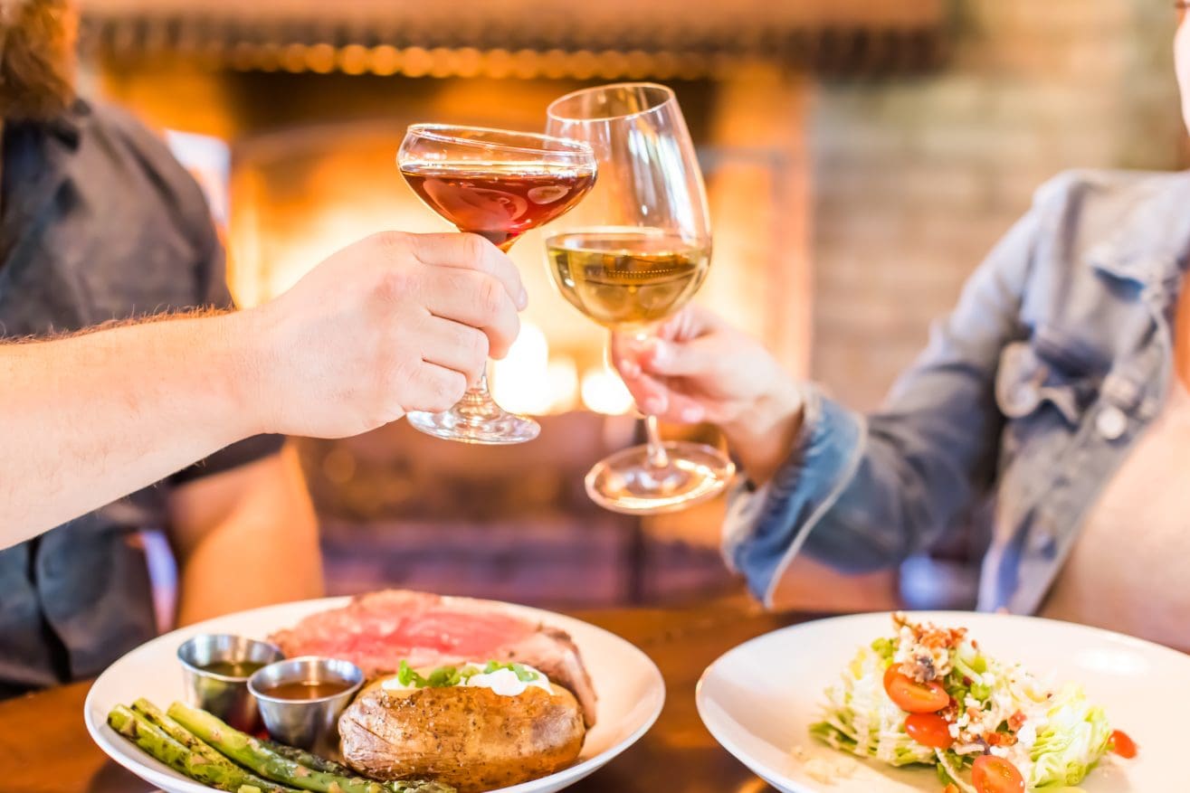 Two people clink wine glasses over a table with plates of steak, baked potato, asparagus, and a wedge salad at a welcoming steakhouse in Paso Robles. A warm fireplace glows in the background, creating a cozy dining atmosphere.