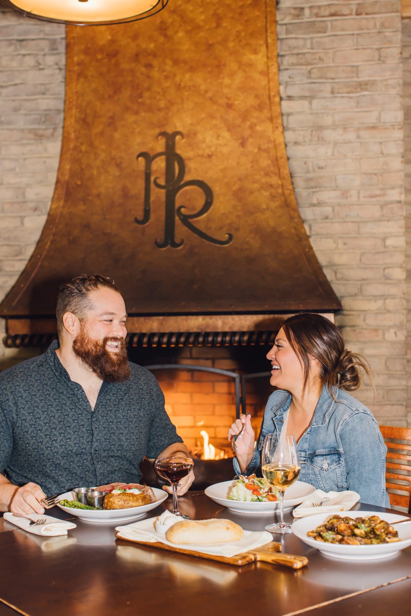 A bearded man and a woman sit at a table in front of a fireplace, enjoying dinner and drinks at a Steakhouse Paso Robles. They are smiling and laughing, with plates of food and glasses of wine on the table.