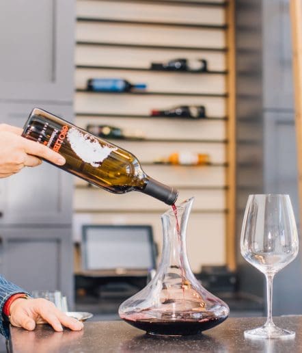 A person pours red wine from a bottle into a glass decanter on a counter, capturing the elegant experience found in many Paso Robles restaurants, with an empty wine glass nearby and shelves of bottles blurred in the background.