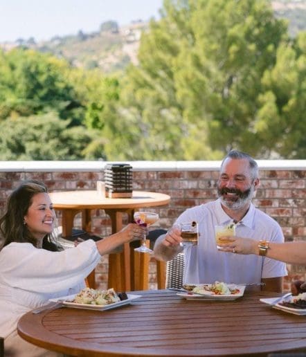Three people sit at a round outdoor table at one of the charming Paso Robles restaurants, smiling and raising drinks in a toast. Plates of food are in front of them, with trees and hills visible in the background.