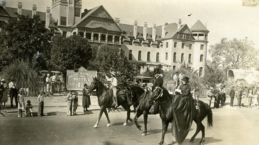A vintage photo shows three people riding horses in front of the historic Paso Robles Hotel. Onlookers, including children, line the sidewalk beneath trees, with the large hotel building in the background.