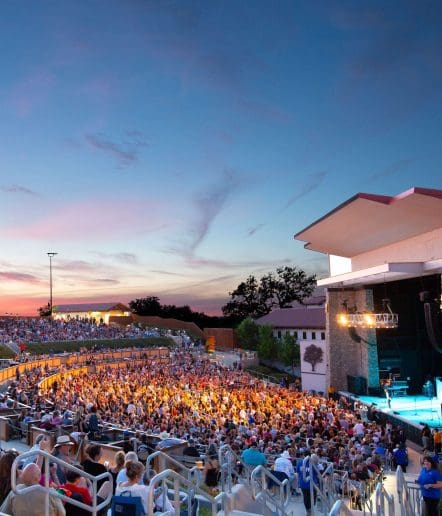 An outdoor amphitheater at sunset hosts a vibrant Paso Robles Events gathering, with a large audience watching a live performance. The colorful sky and stage lights create a magical atmosphere as the crowd enjoys the show in tiered rows.