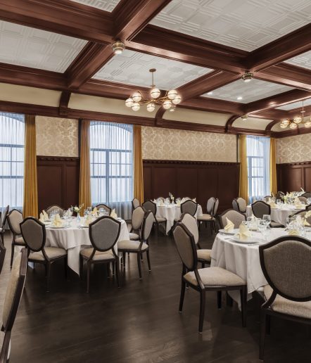 Elegant dining room with round tables covered in white cloths, set with glassware and napkins, surrounded by upholstered chairs. The room features wood paneling, large windows with curtains, and vintage ceiling lights.