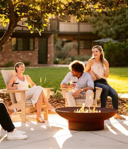 Three adults sit around a fire pit outdoors, laughing and holding glasses of wine, with sunlight filtering through trees and a building visible in the background.