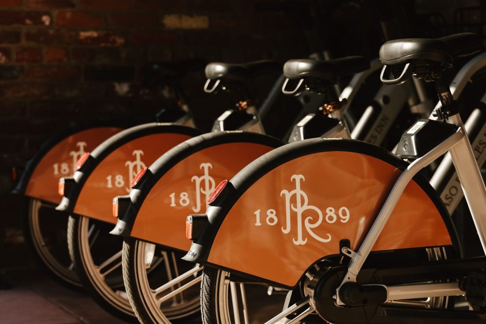Four orange rental bikes are lined up next to each other, each displaying “J R 1889” in white on the rear wheel covers. The bikes are parked indoors, with a brick wall in the background.