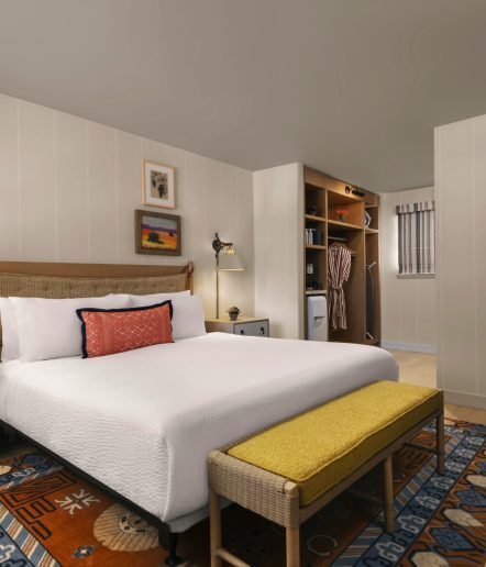 A cozy bedroom with a neatly made bed, a yellow cushioned bench at the foot, an open closet with a robe, framed art on the walls, a colorful rug, and a small window letting in natural light.