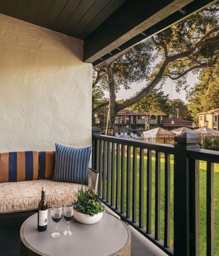 A cozy outdoor patio with a striped cushioned bench, blue pillow, round table with a bottle of wine, two glasses, and a plant. The patio overlooks a green lawn and trees with buildings in the background.