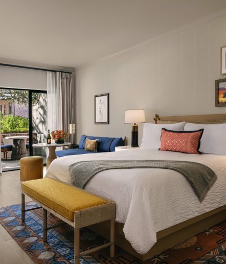 A modern hotel room with a large bed, gold bench, blue sofa, artwork on the walls, a nightstand with a lamp, and glass doors opening to a patio with outdoor seating and greenery visible outside.