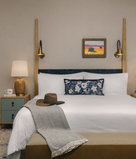 A neatly made bed with white linens, a floral pillow, and a brown hat and gray throw on the end. Two modern wall lamps, a nightstand with a lamp and mug, and a landscape painting hang above the bed.